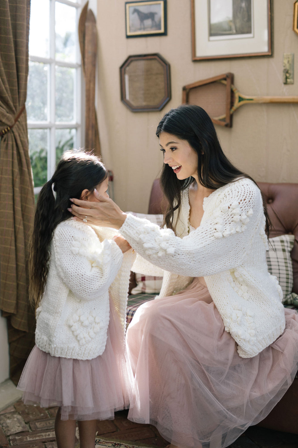 Two women in white knit cardigans and blush pink tulle skirts, smiling and embracing in a cozy home setting.