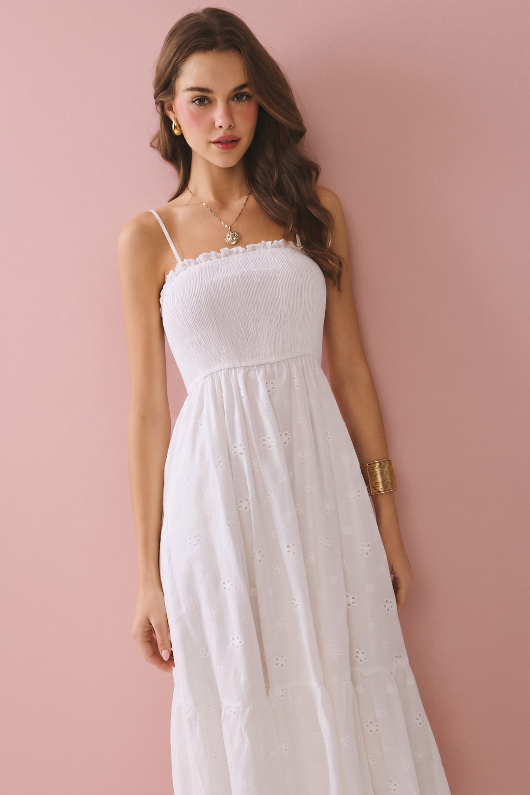 A woman wearing a white spaghetti-strap midi dress with a smocked bodice and embroidered details, posing against a solid dusty pink background.