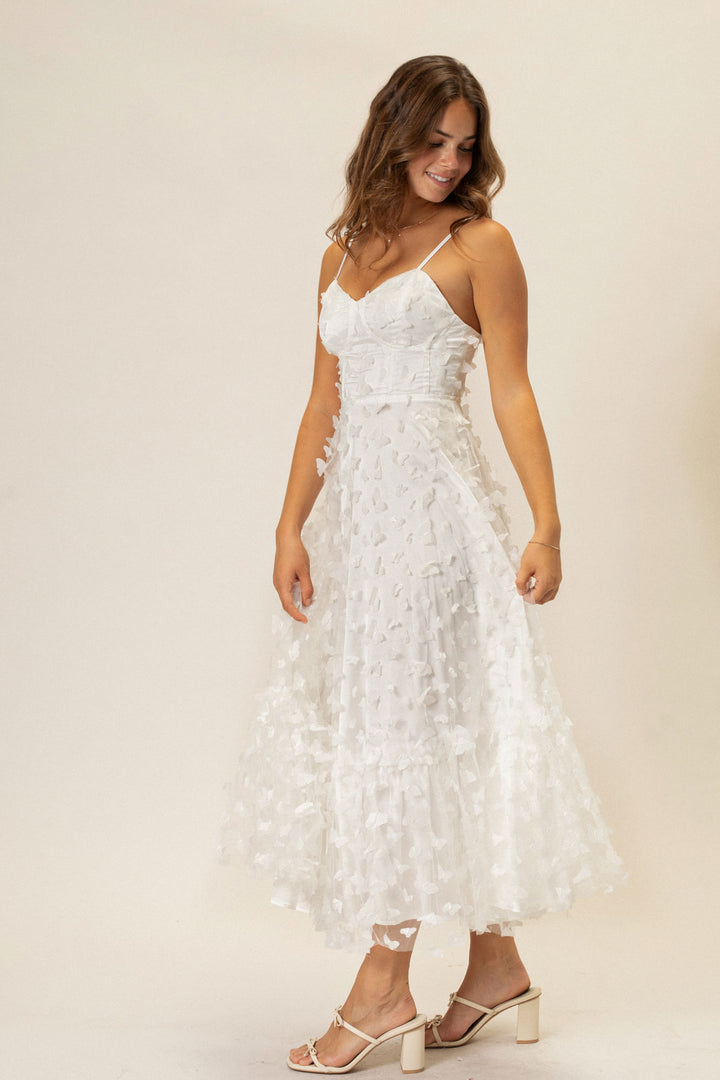 A white knee-length dress with intricate butterfly-patterned mesh overlay, featuring spaghetti straps and a flowy silhouette.