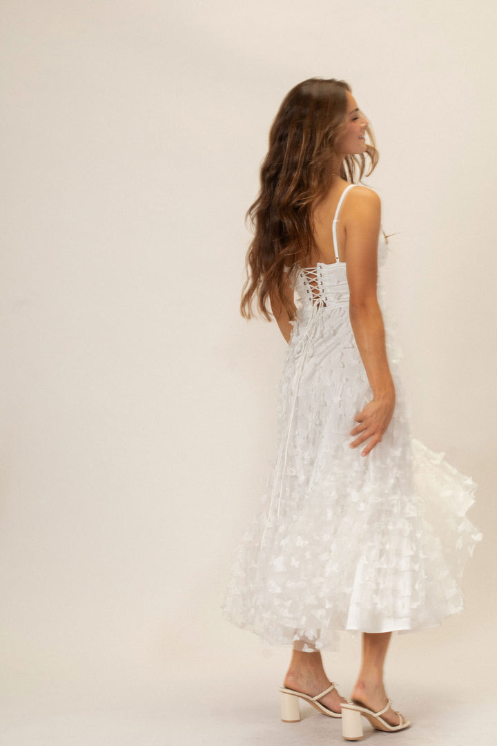 A white, floor-length dress with a lace-up back and fluttery, butterfly-like mesh details cascading down the skirt, worn by a woman with long, wavy brown hair.
