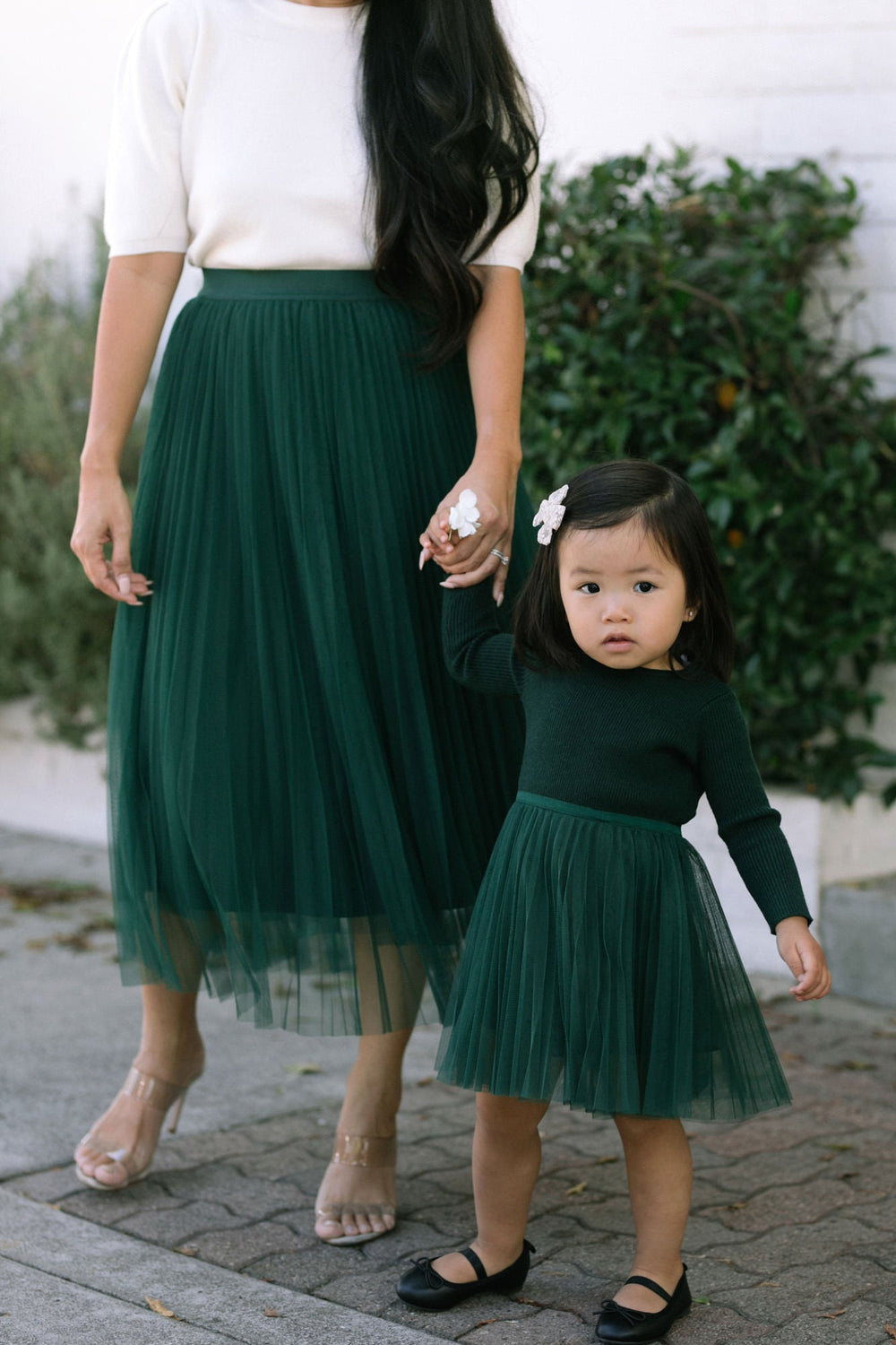 A young girl in a green pleated tulle dress holding the hand of a woman wearing a white top and green pleated midi skirt, standing in a garden setting.