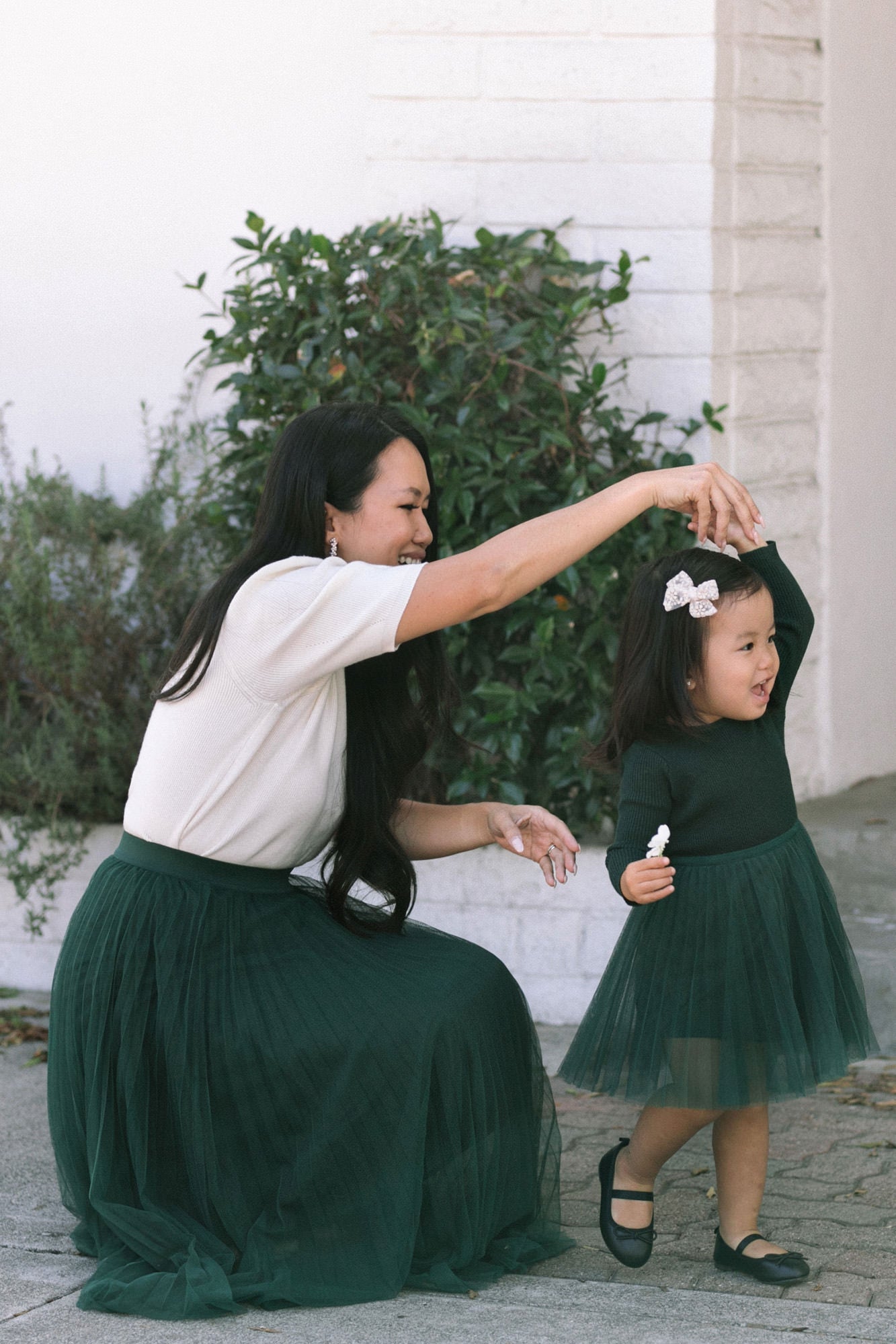 A woman in a white top and a young girl in a green tulle dress stand together, with the woman's arm around the girl, in a garden setting.