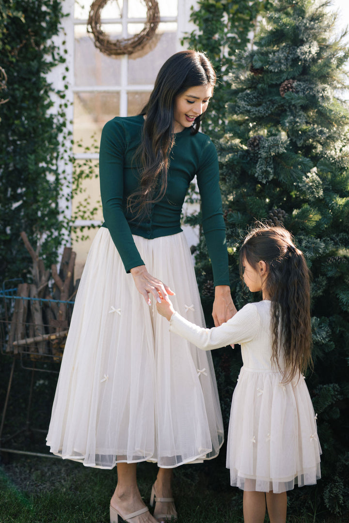 A woman in a green top and a flowing white tulle skirt with bows, standing with a young girl in a white dress, against a Christmas tree backdrop.