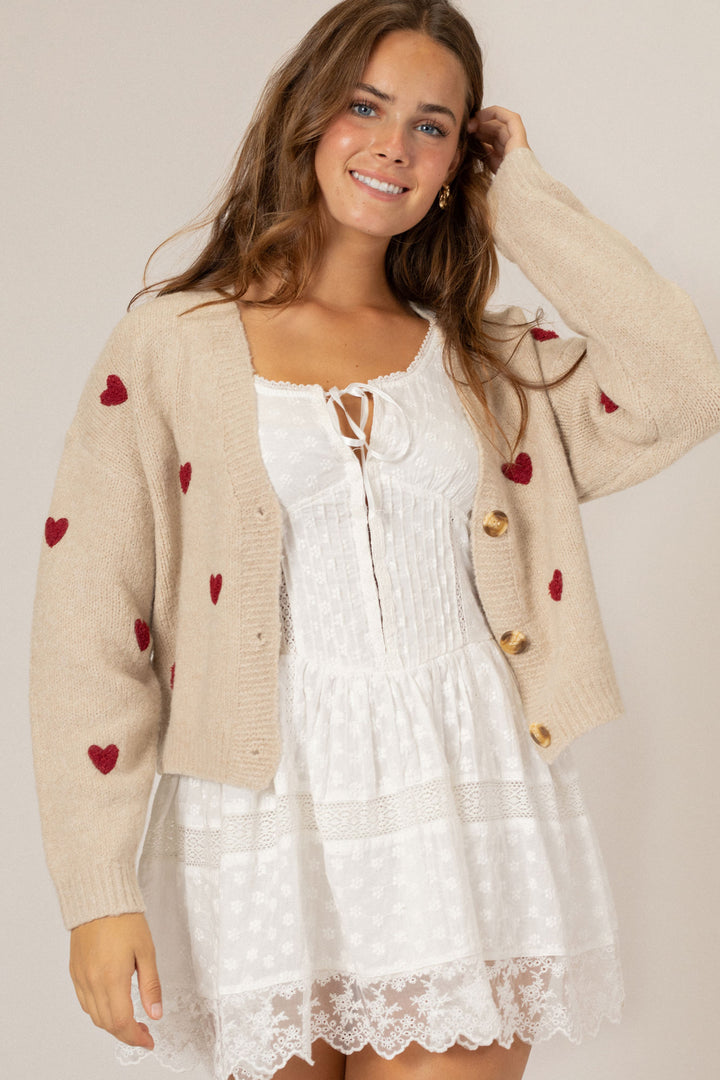 A beige, knit cardigan with red heart-shaped designs, featuring a white lace-trimmed dress underneath, modeled by a smiling woman.