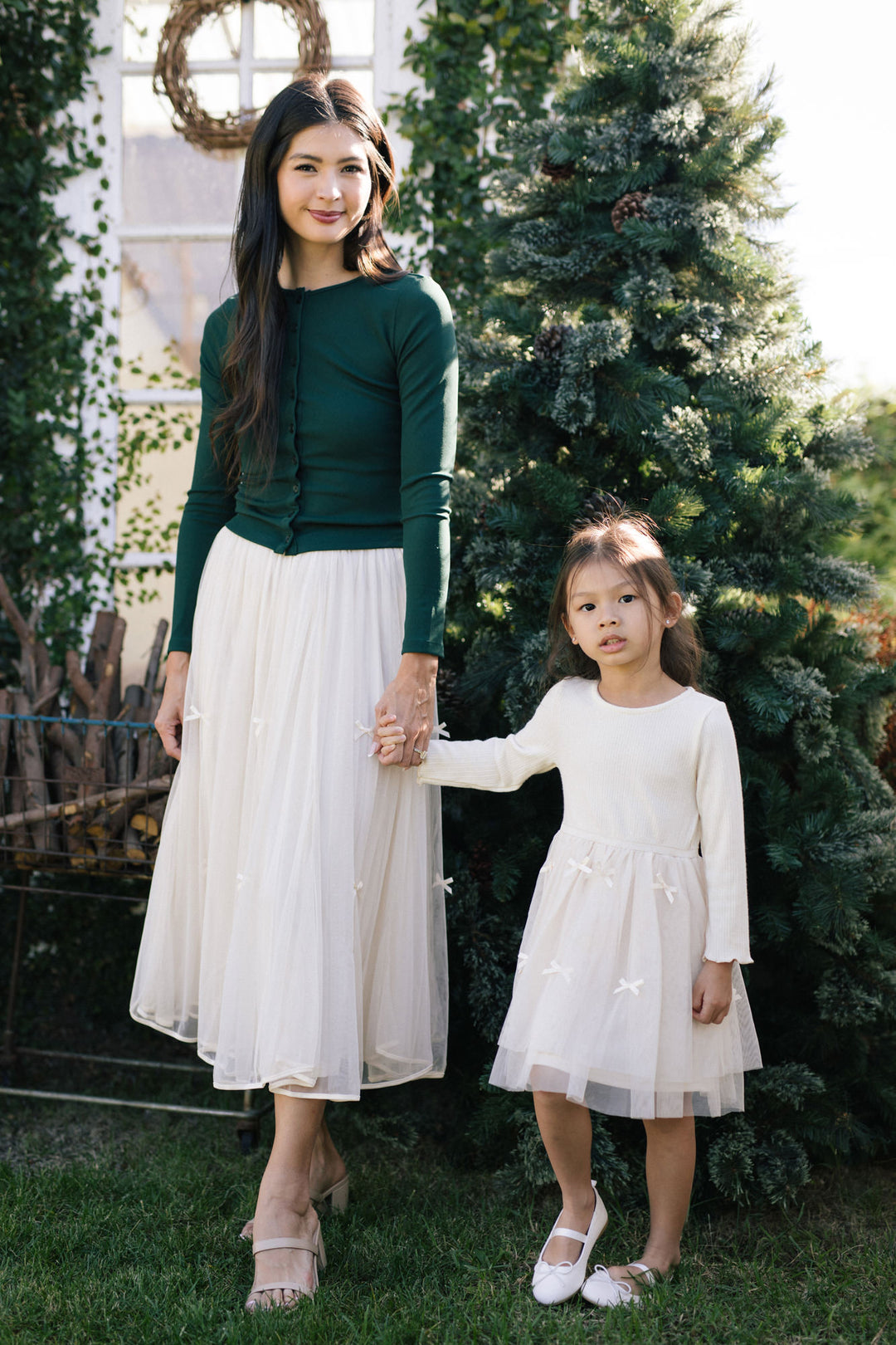 A woman in a green top and a young girl in a white tulle dress are standing together in front of a Christmas tree, holding hands and smiling.