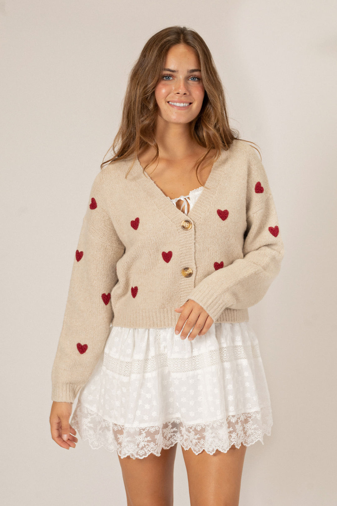 A beige knit cardigan with red heart patterns, featuring gold buttons and a flouncy white lace mini dress underneath.