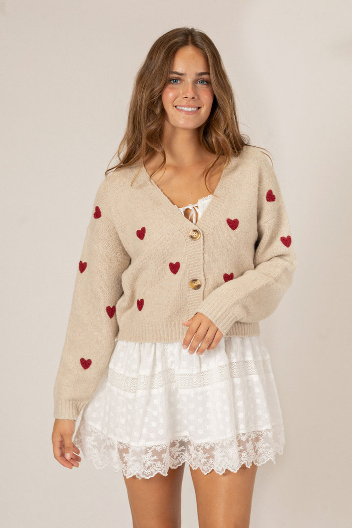 A beige knit cardigan with red heart patterns, featuring gold buttons and a flouncy white lace mini dress underneath.