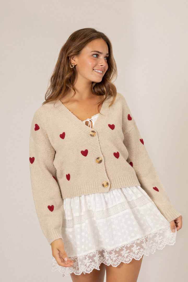 A beige knit cardigan with scattered burgundy heart-shaped embroidered details, featuring a peplum silhouette and gold button closure.