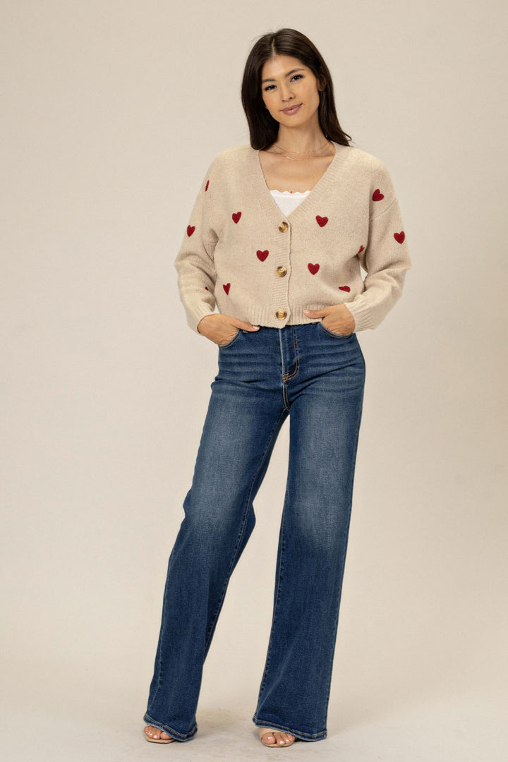 A cream-colored knit cardigan with red heart designs, buttoned down the front, worn with high-waisted blue jeans by a smiling model.