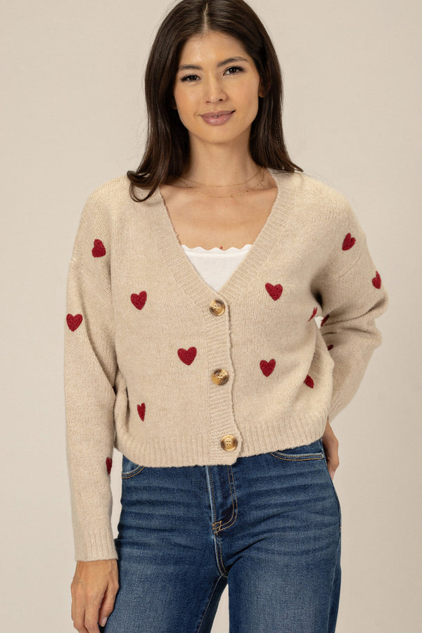 Beige knitted cardigan with red heart embroidery, featuring a V-neck and button closure, worn by a smiling young woman with long dark hair.