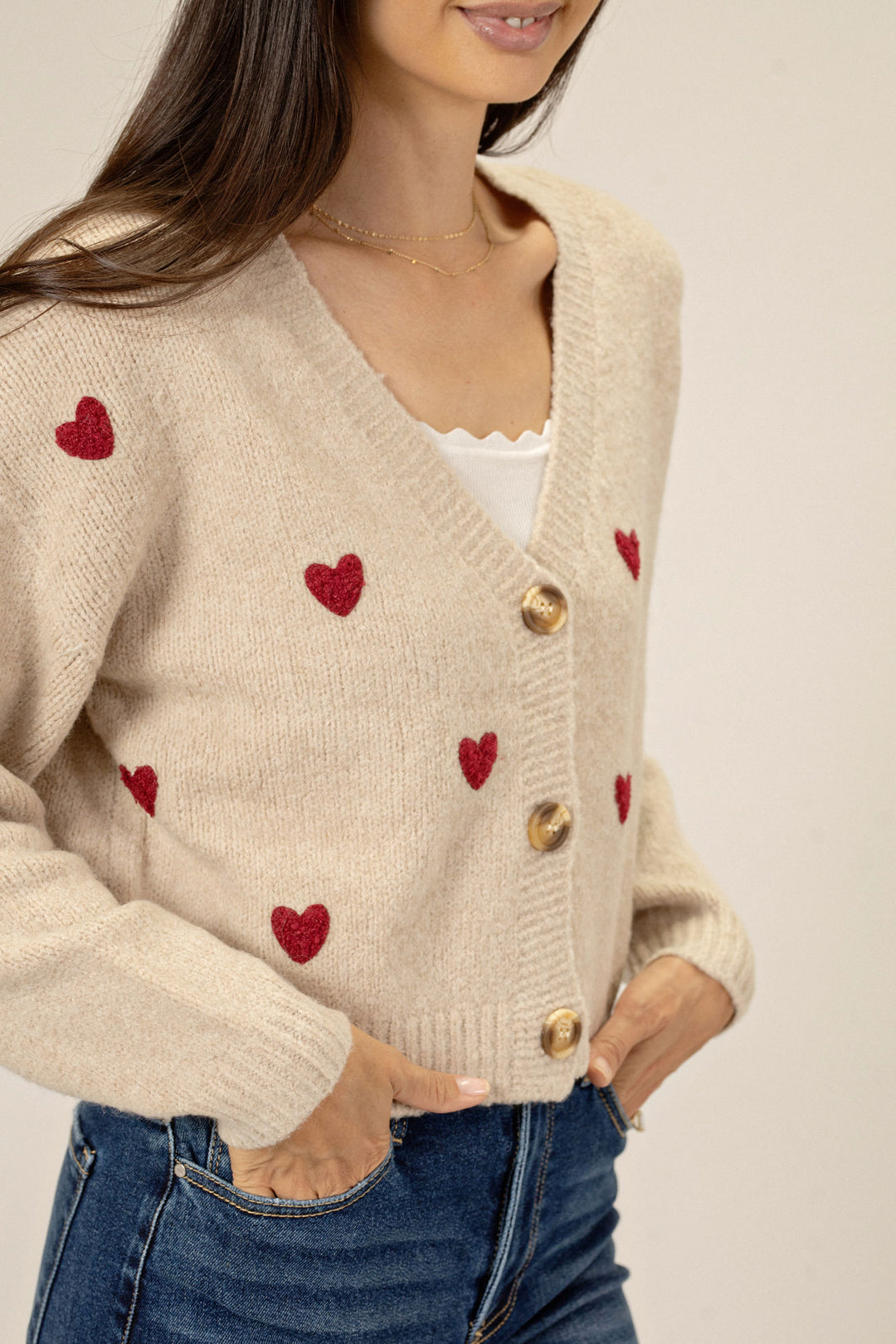 Beige knit cardigan with red heart patterns, buttoned closure, relaxed fit, perfect for casual or dressed-up looks.
