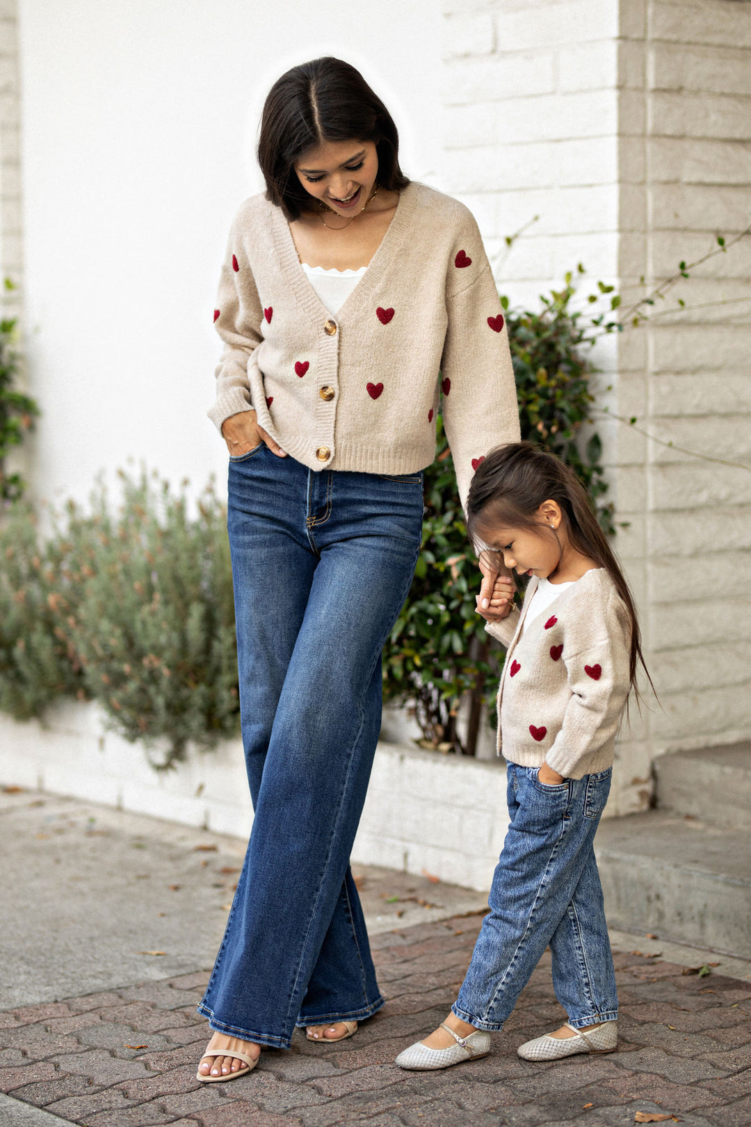 A smiling woman wearing a beige cardigan with red heart patterns, standing next to a young girl wearing a matching heart-patterned sweater, both with jeans, in a backyard setting.