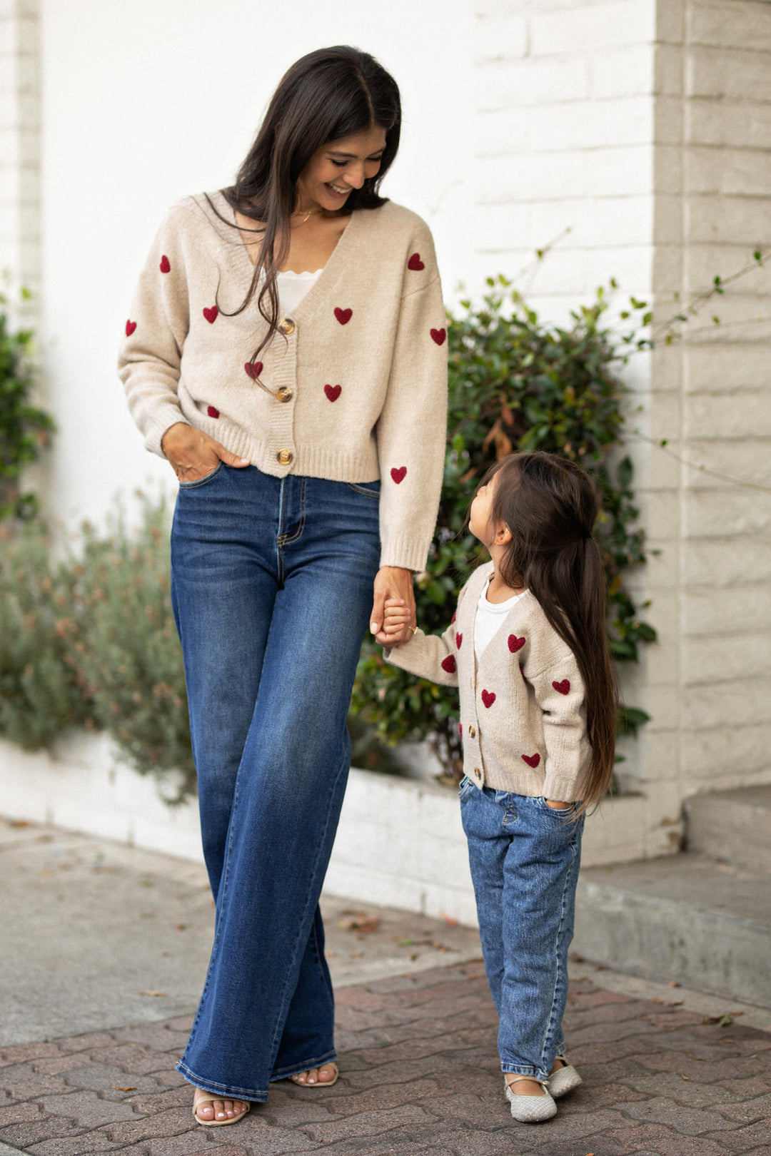 A woman and child wearing matching sweaters with red heart designs, smiling and holding hands while standing outdoors.