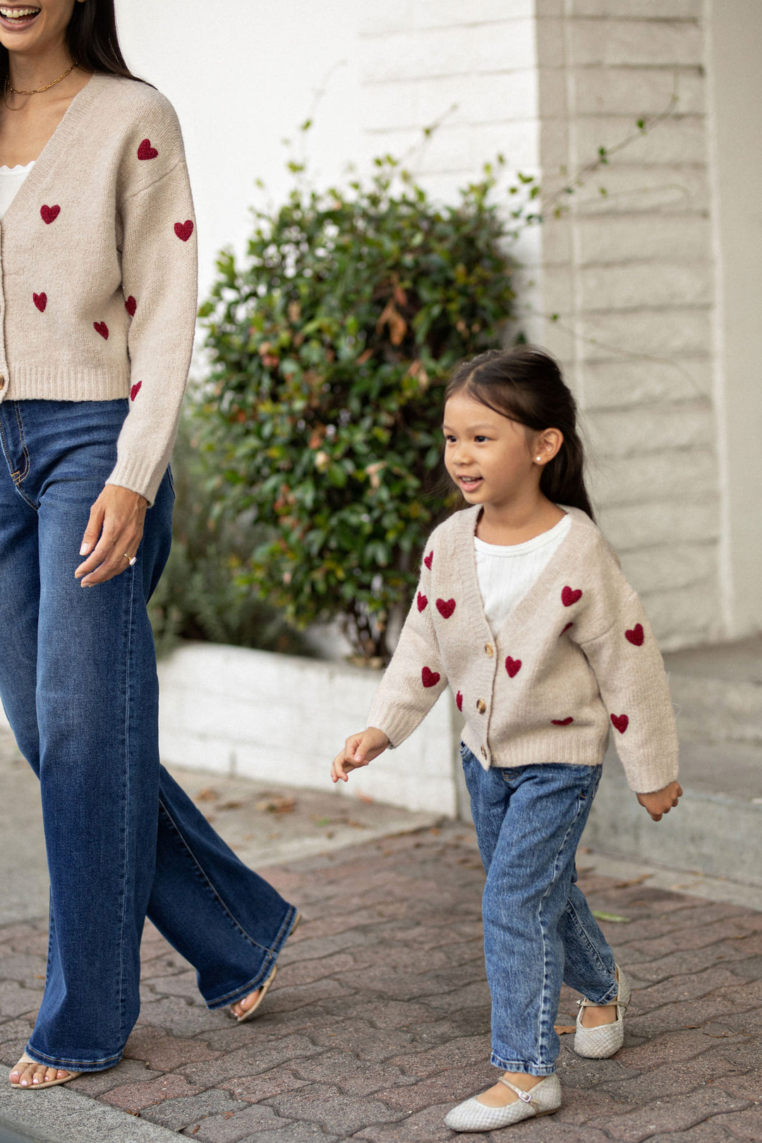 A young girl joyfully smiles while wearing a beige cardigan with red heart designs, standing next to an adult woman in a similar heart-patterned sweater in an outdoor setting.