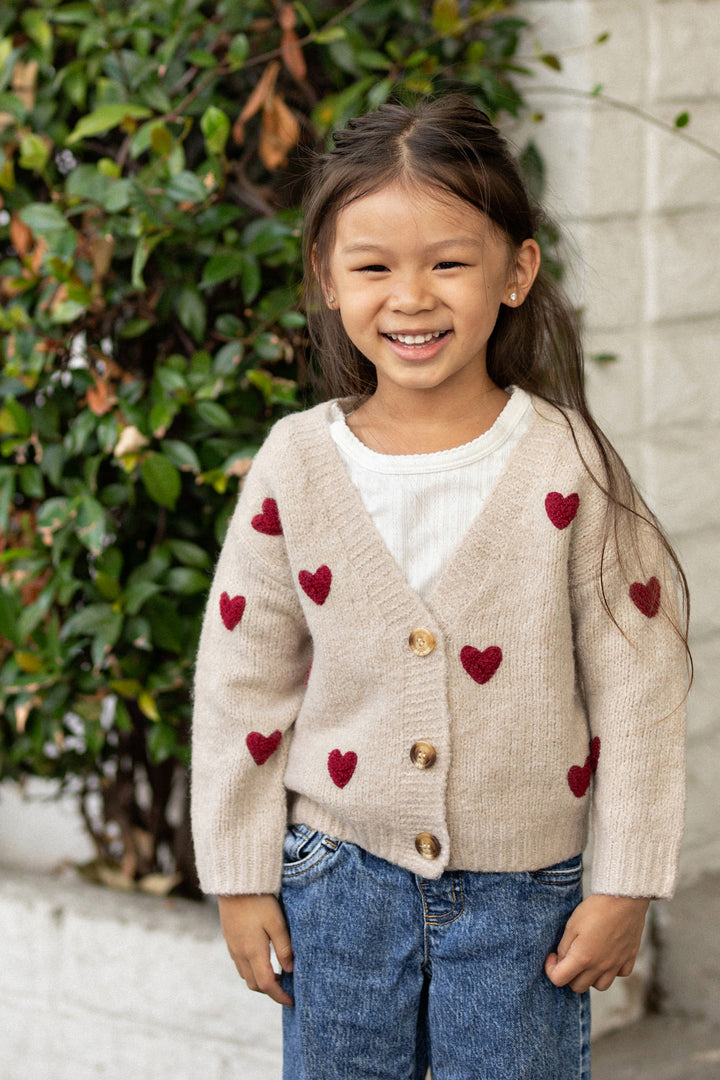A young girl smiles brightly, wearing a beige cardigan with red heart patterns, paired with denim jeans, against a backdrop of lush greenery.