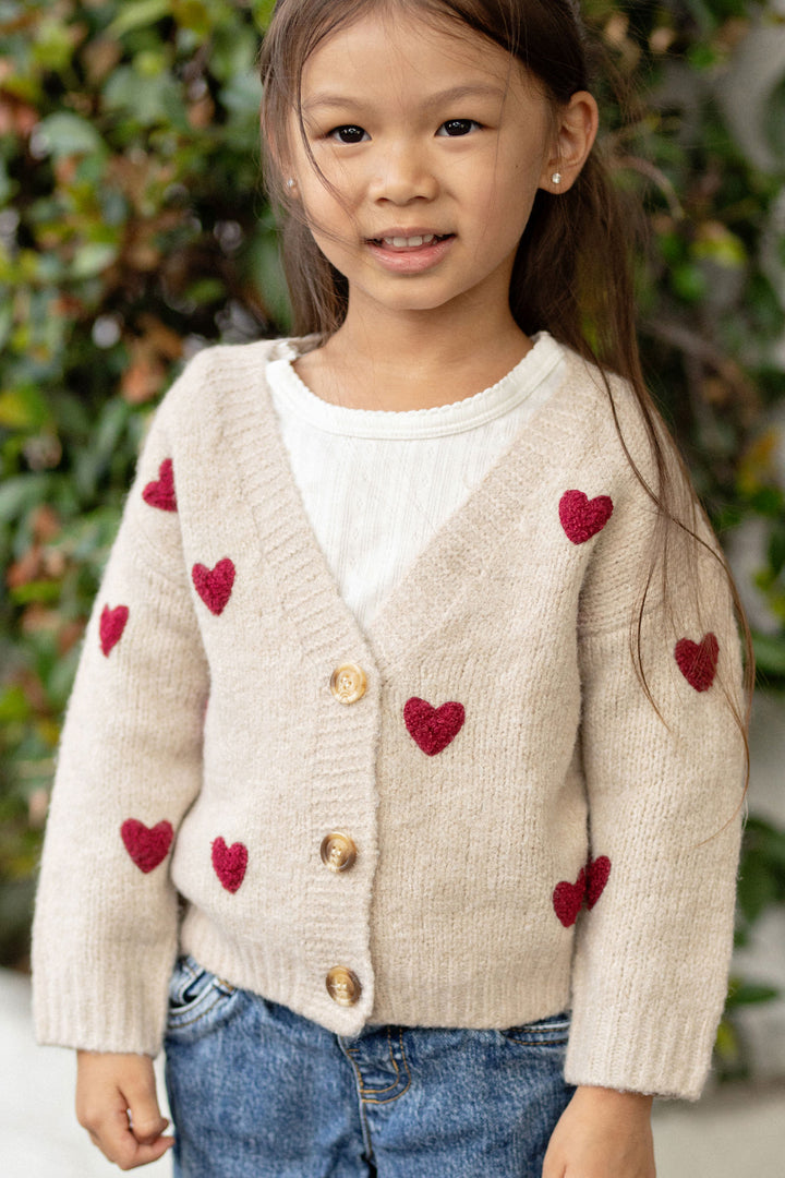 A young girl wearing a cream-colored cardigan sweater with red heart patterns, posing outdoors against a natural backdrop.