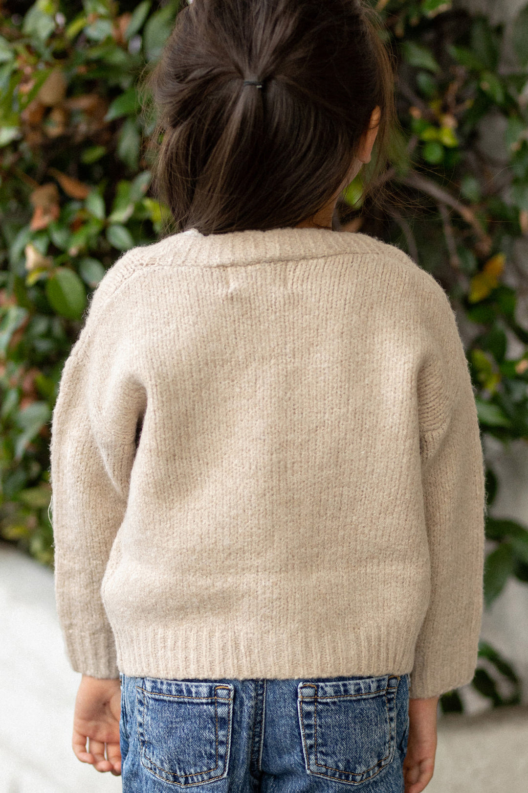 A young girl wearing a cozy, cream-colored knitted cardigan with an asymmetrical hemline, standing against a natural, green backdrop.
