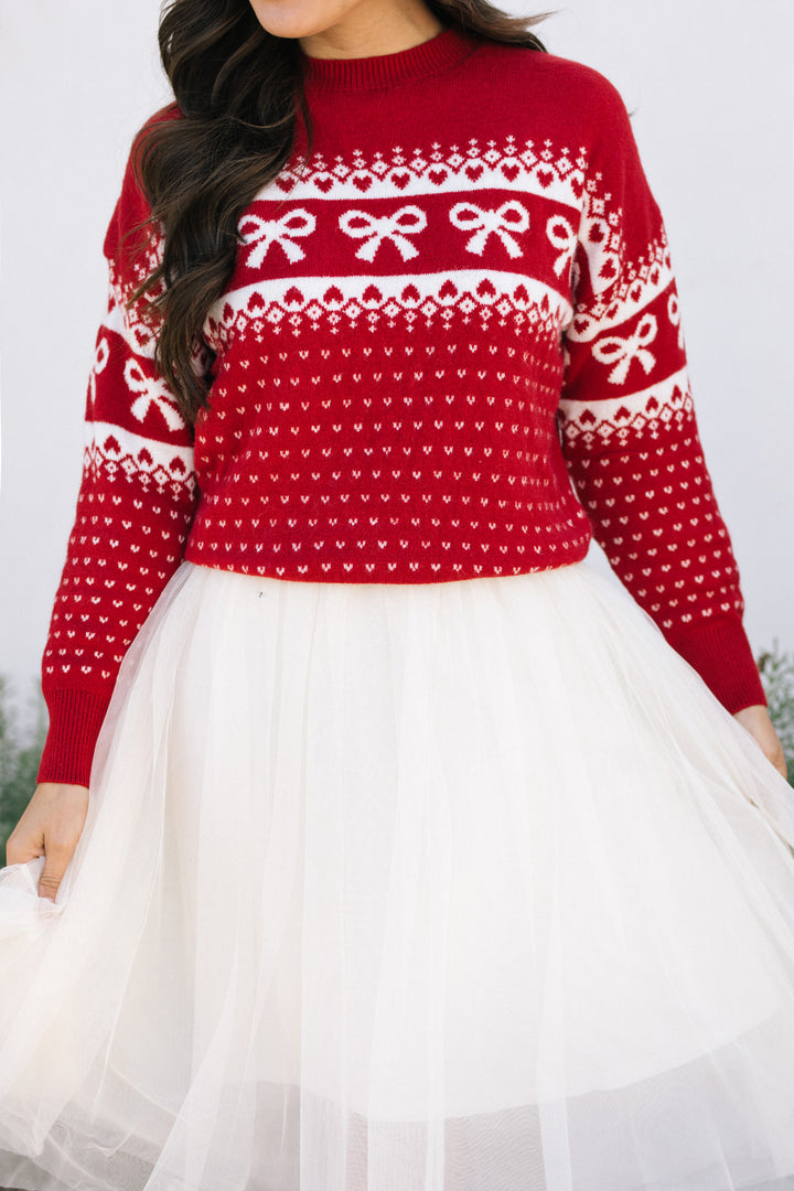 A vibrant red knit sweater with a repeating pattern of white bows and hearts, worn with a flowing white skirt.