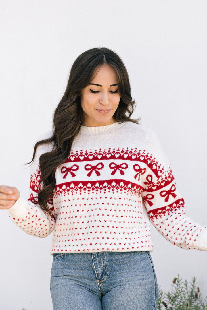 A cozy, festive red and white knit sweater with a repeating bow pattern, worn by a young woman with long, wavy dark hair.