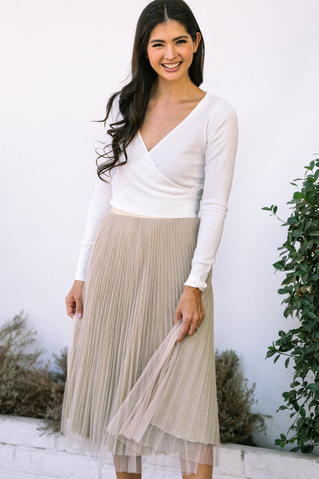 White blouse with a deep V-neck, long sleeves, and beige pleated midi skirt with a tulle underlay.
