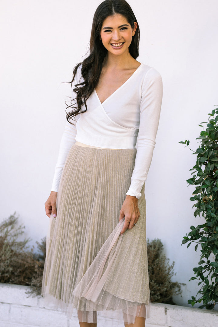 White blouse with a deep V-neck, long sleeves, and beige pleated midi skirt with a tulle underlay.