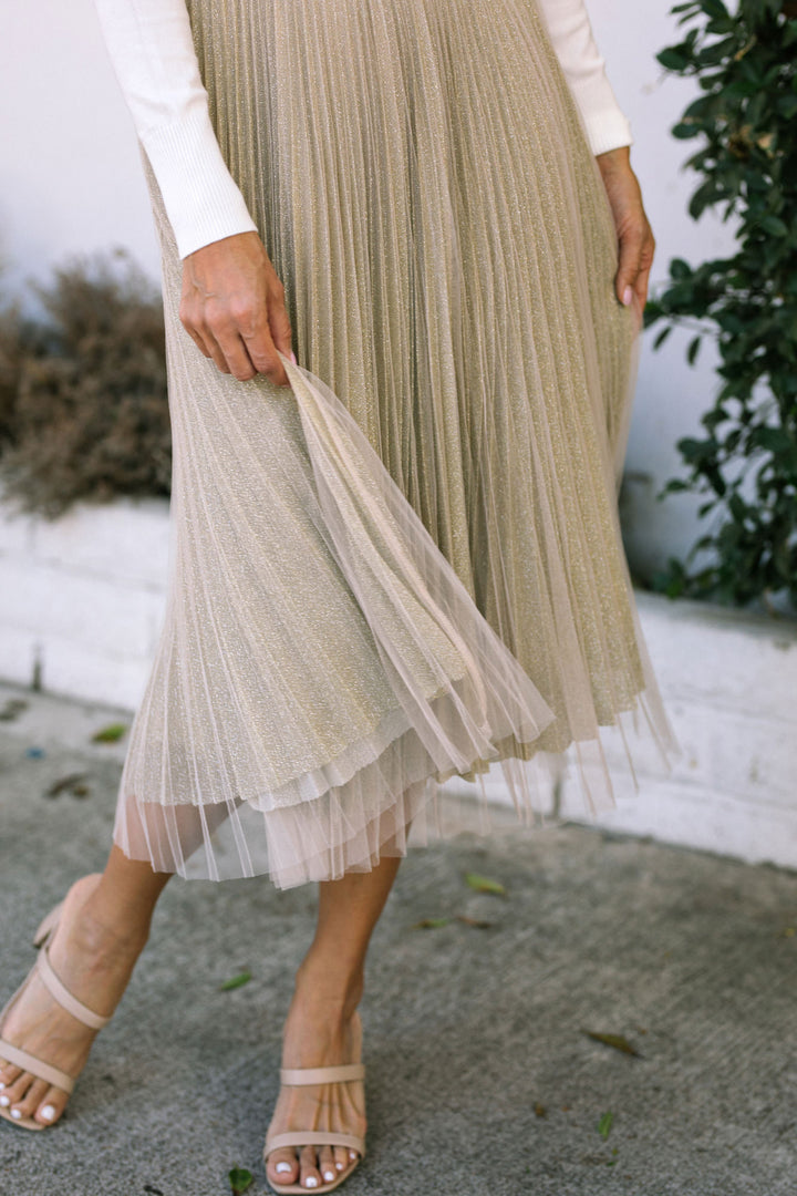 Gold pleated midi skirt with sheer mesh overlay, paired with neutral strappy sandals.