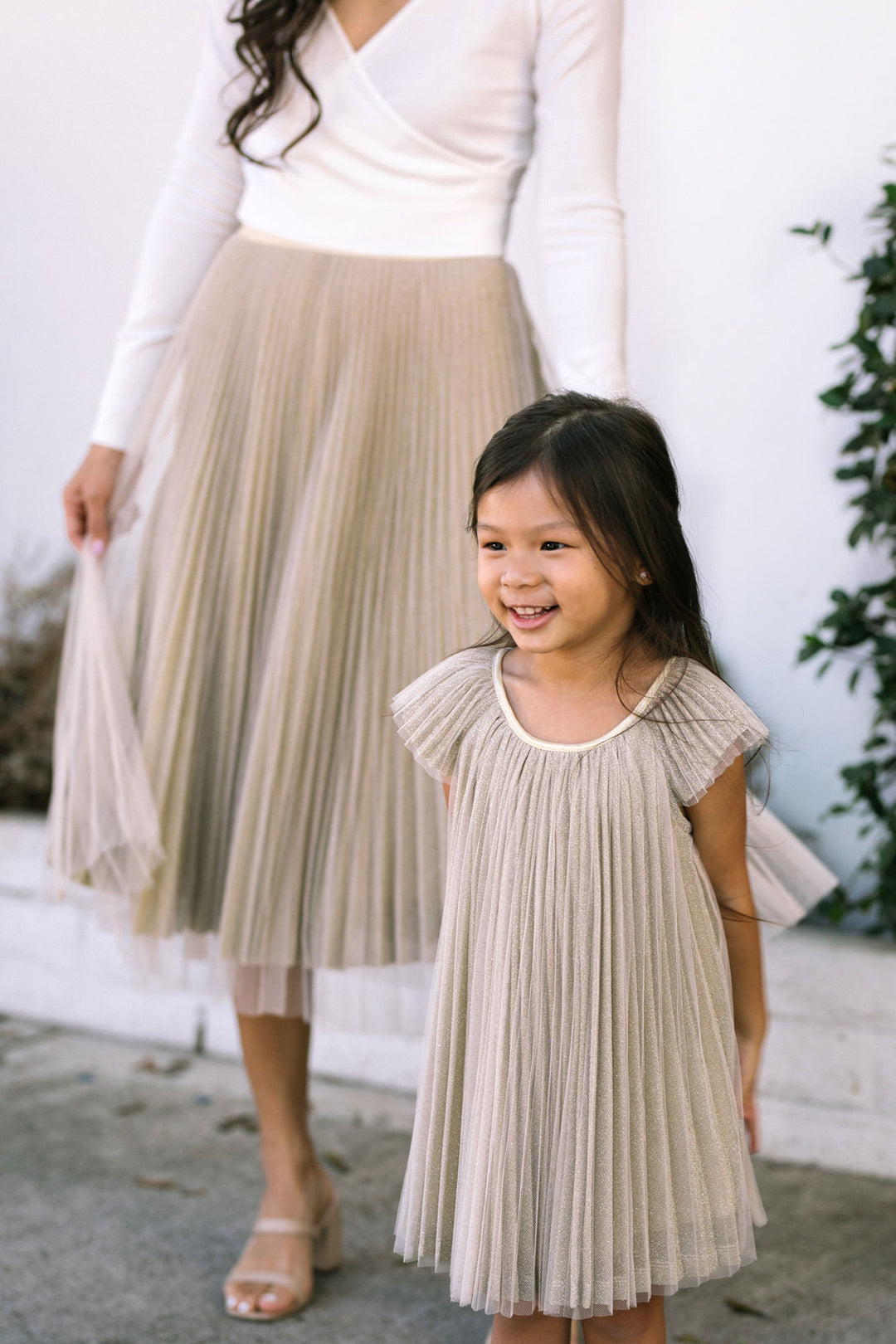 A young girl wearing a pleated dress with a capped sleeve and princess waist in a beige/tan color, smiling happily.
