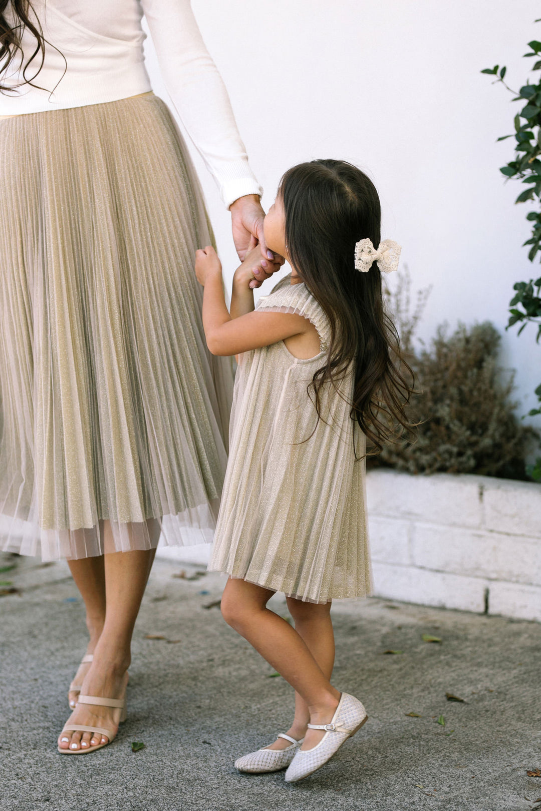 A young girl in a pleated, cream-colored dress with a floral hair accessory, walking with an adult in a neutral background.