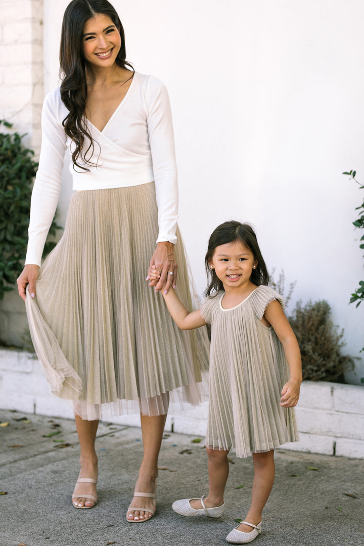 A smiling young girl wearing a pleated beige dress stands next to a woman in a white shirt and beige pleated midi skirt, holding hands.