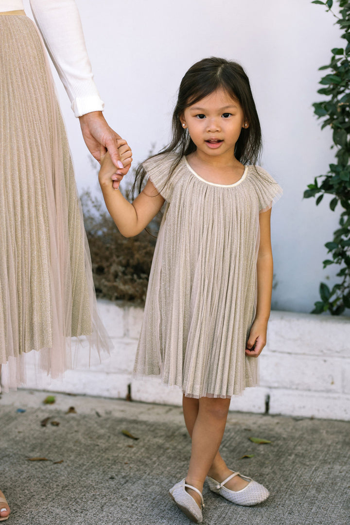 A young girl wearing a light beige pleated dress with short sleeves, standing while holding an adult's hand.