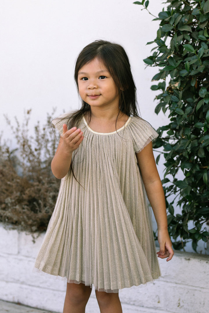 A young girl in a beige, pleated dress with short sleeves, smiling against a backdrop of green leaves.