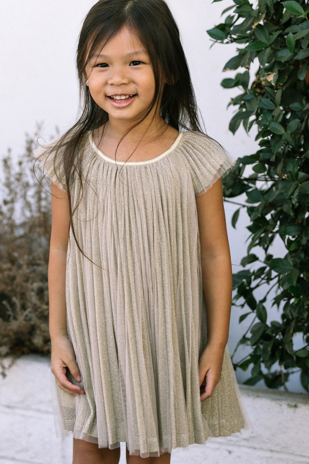 A young girl in a beige pleated dress with short sleeves, standing in front of greenery and smiling brightly.