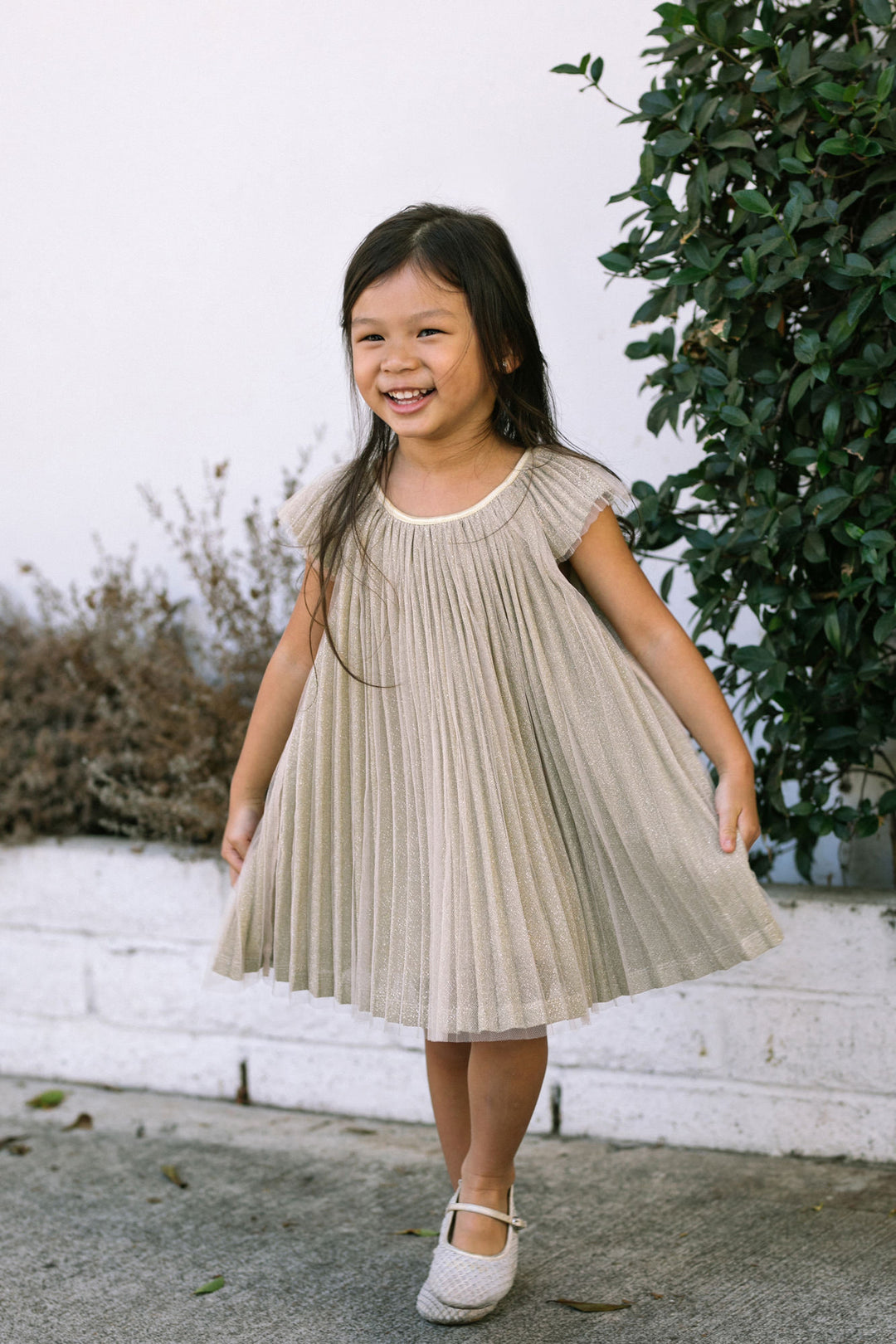 A young girl wearing a beige, pleated dress with short sleeves, standing outdoors amid lush greenery and smiling brightly.