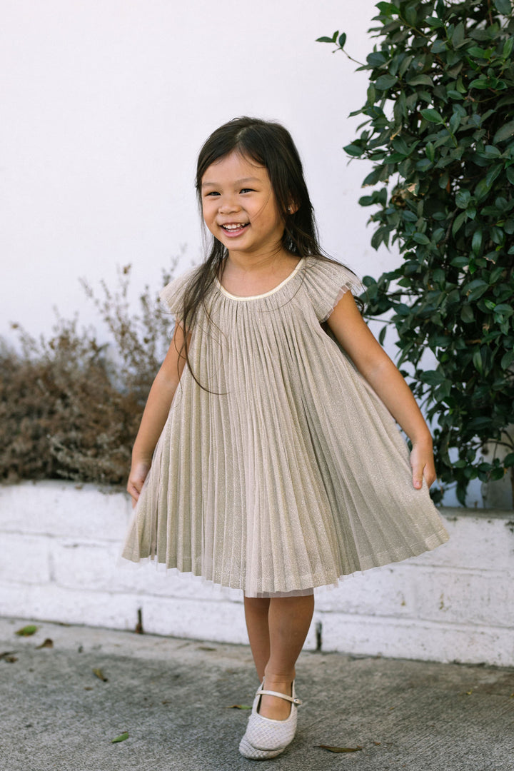 A young girl wearing a beige, pleated dress with short sleeves, standing outdoors amid lush greenery and smiling brightly.