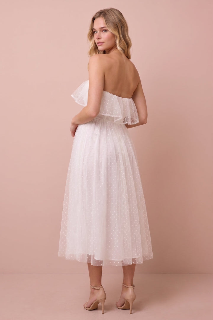 A woman modeling a strapless white tulle midi dress with a ruffled off-the-shoulder neckline, shown from the back against a solid dusty pink studio backdrop.