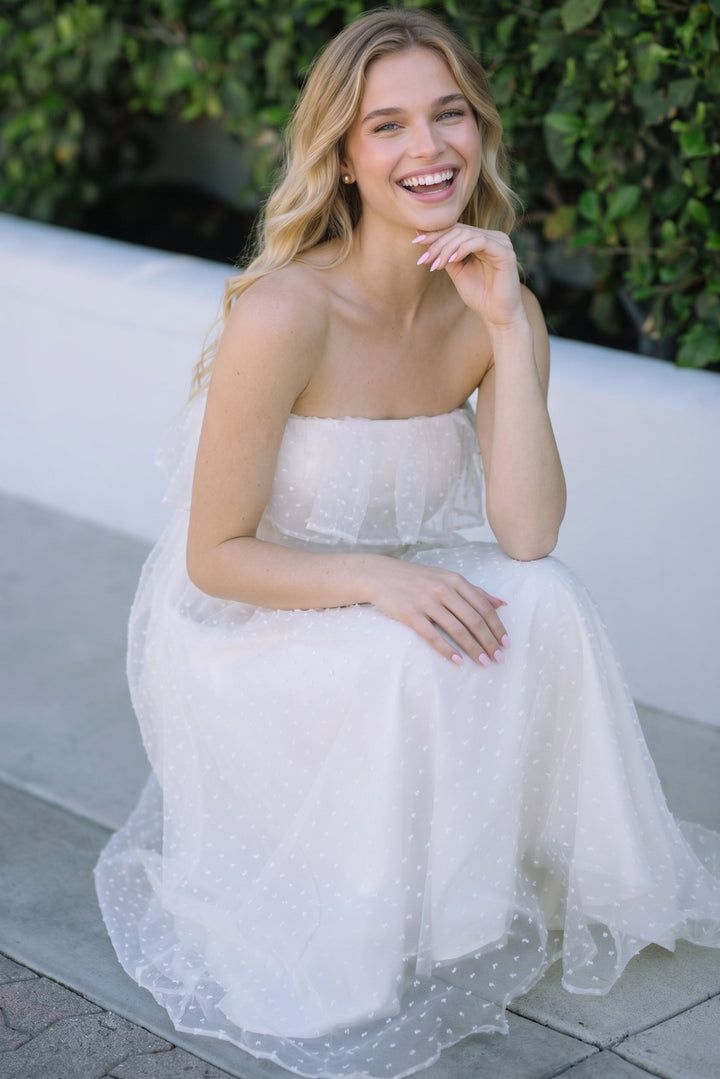 A woman wearing a strapless white tulle dress with a sheer overlay and small polka dots, sitting outdoors against a white wall and green foliage background.