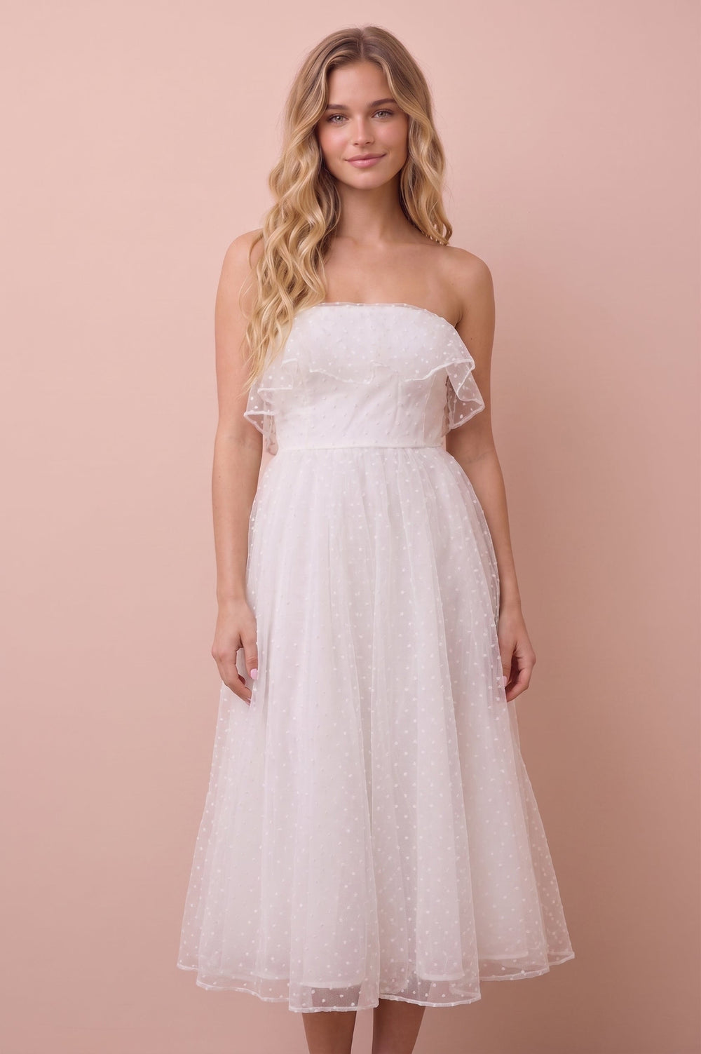 A woman modeling a strapless white tulle midi dress with ruffled shoulder details against a solid pale pink studio background.