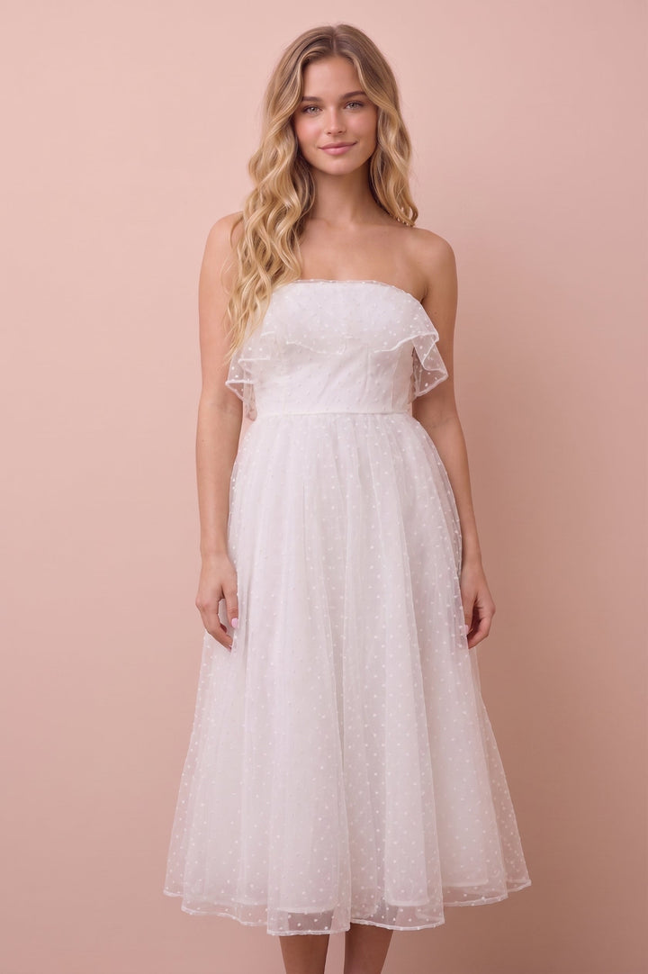 A woman modeling a strapless white tulle midi dress with ruffled shoulder details against a solid pale pink studio background.