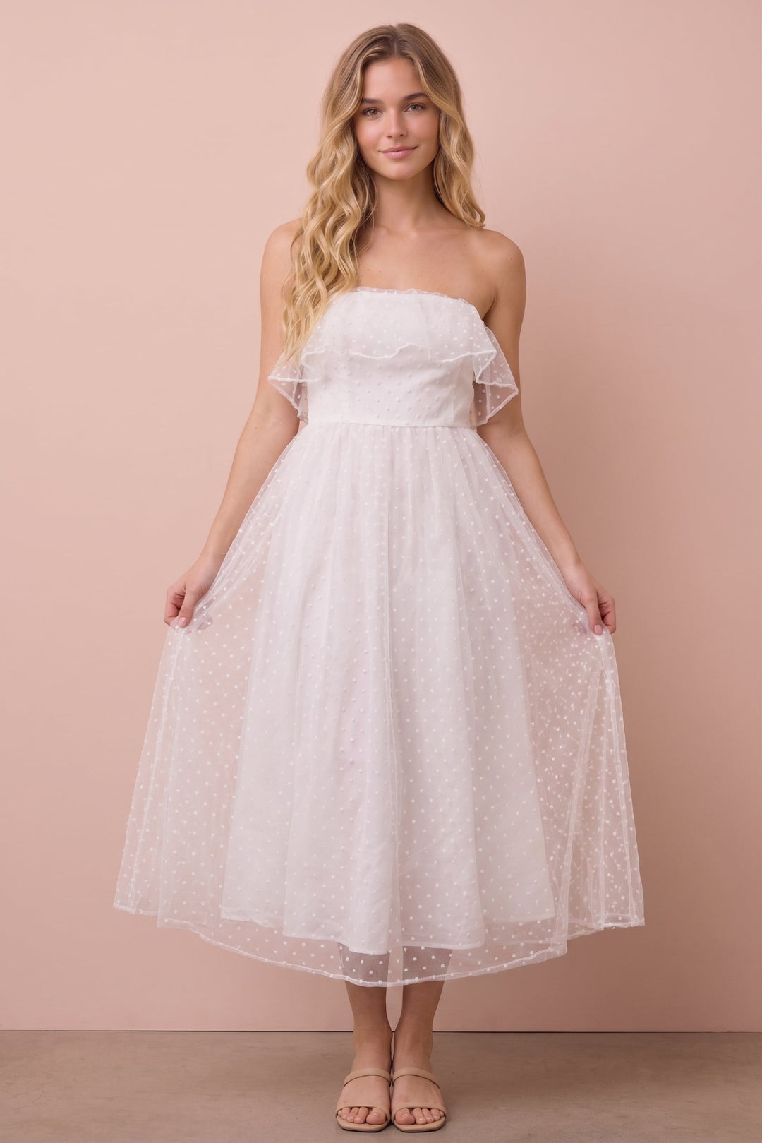 A woman modeling a strapless white tulle midi dress with ruffle details on the shoulders and a flared skirt, standing against a solid dusty pink background.