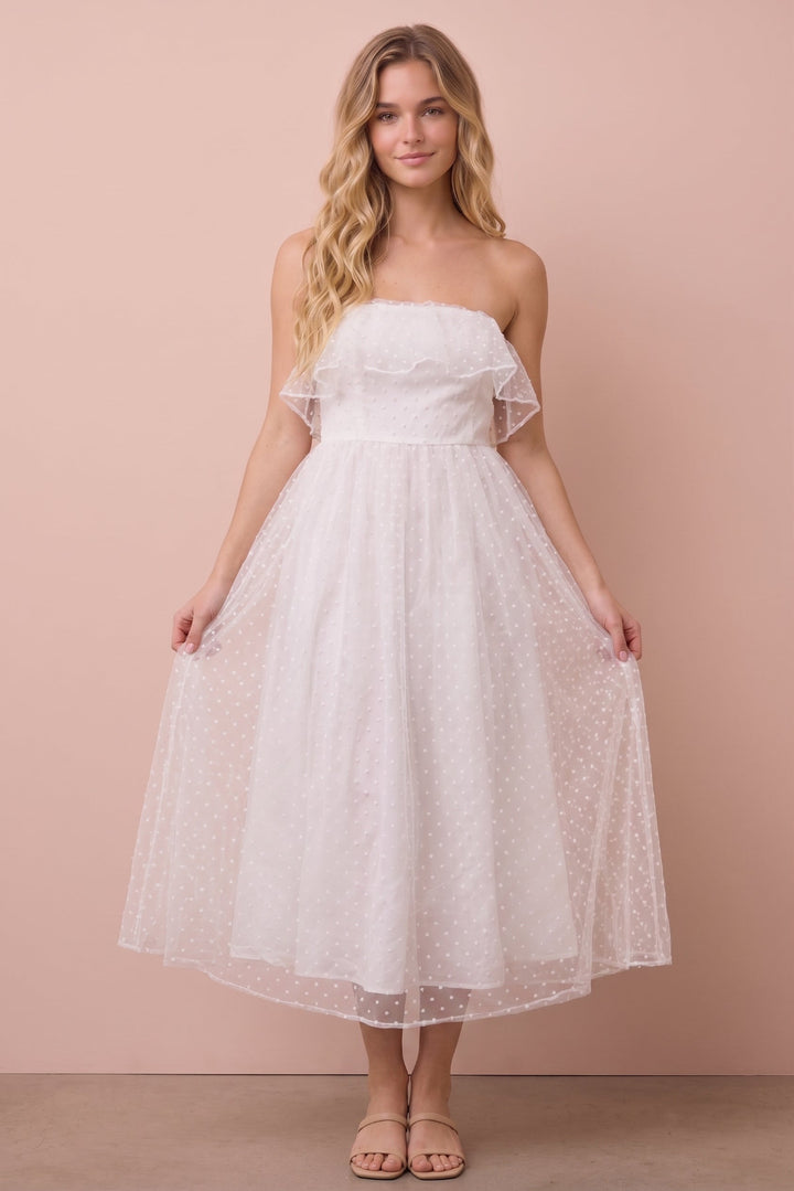 A woman modeling a strapless white tulle midi dress with ruffle details on the shoulders and a flared skirt, standing against a solid dusty pink background.