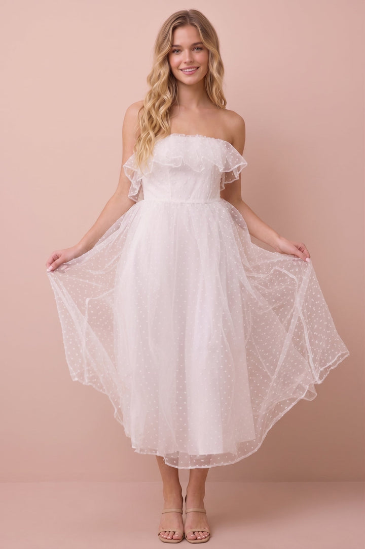 A woman modeling a white off-the-shoulder midi dress with a ruffled neckline and a flared tulle skirt, standing against a solid pale pink studio backdrop.