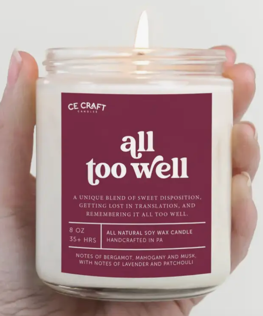 A burgundy-colored glass candle jar with the text "all too well" and a description of its unique blend of scents.