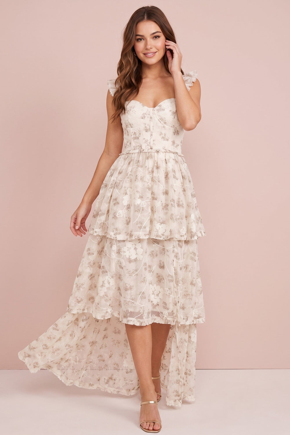 A woman models a sleeveless high-low dress with a sweetheart neckline, a tiered ruffled skirt, and a floral lace overlay, set against a solid pale pink studio background.