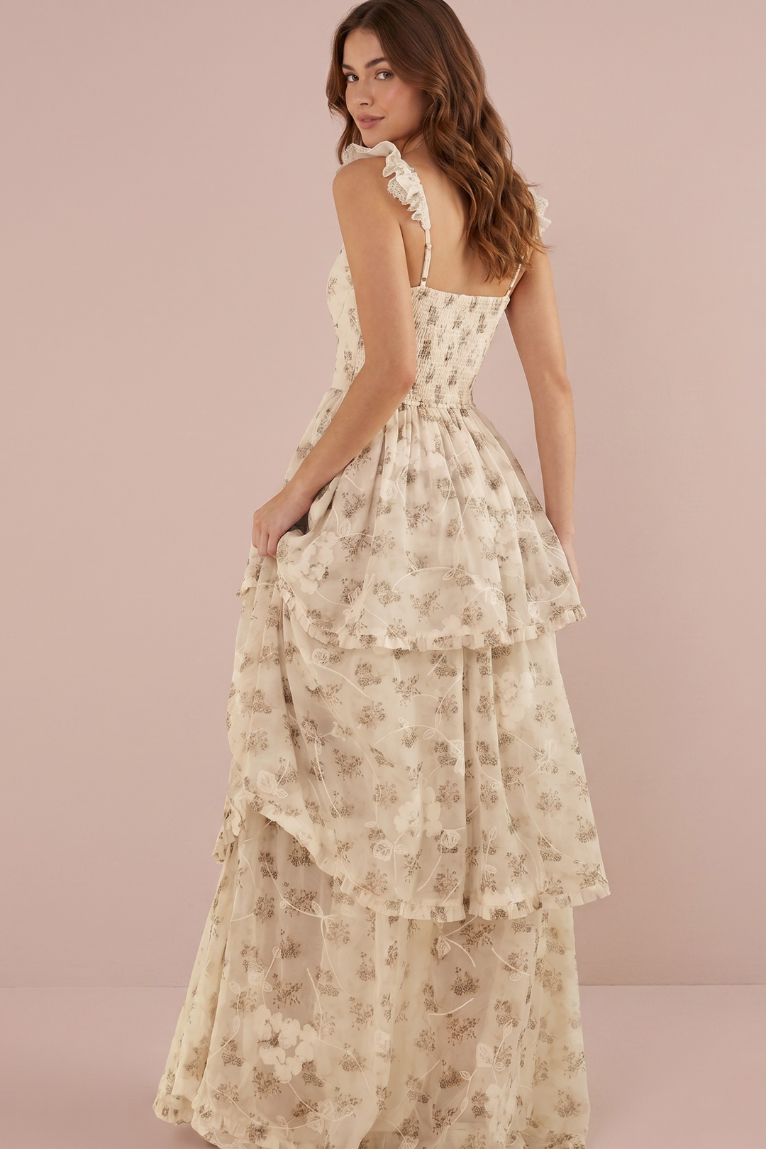 A woman modeling a cream-colored, tiered maxi dress with a delicate beige floral pattern, shown from a three-quarters back view against a solid light pink studio backdrop.