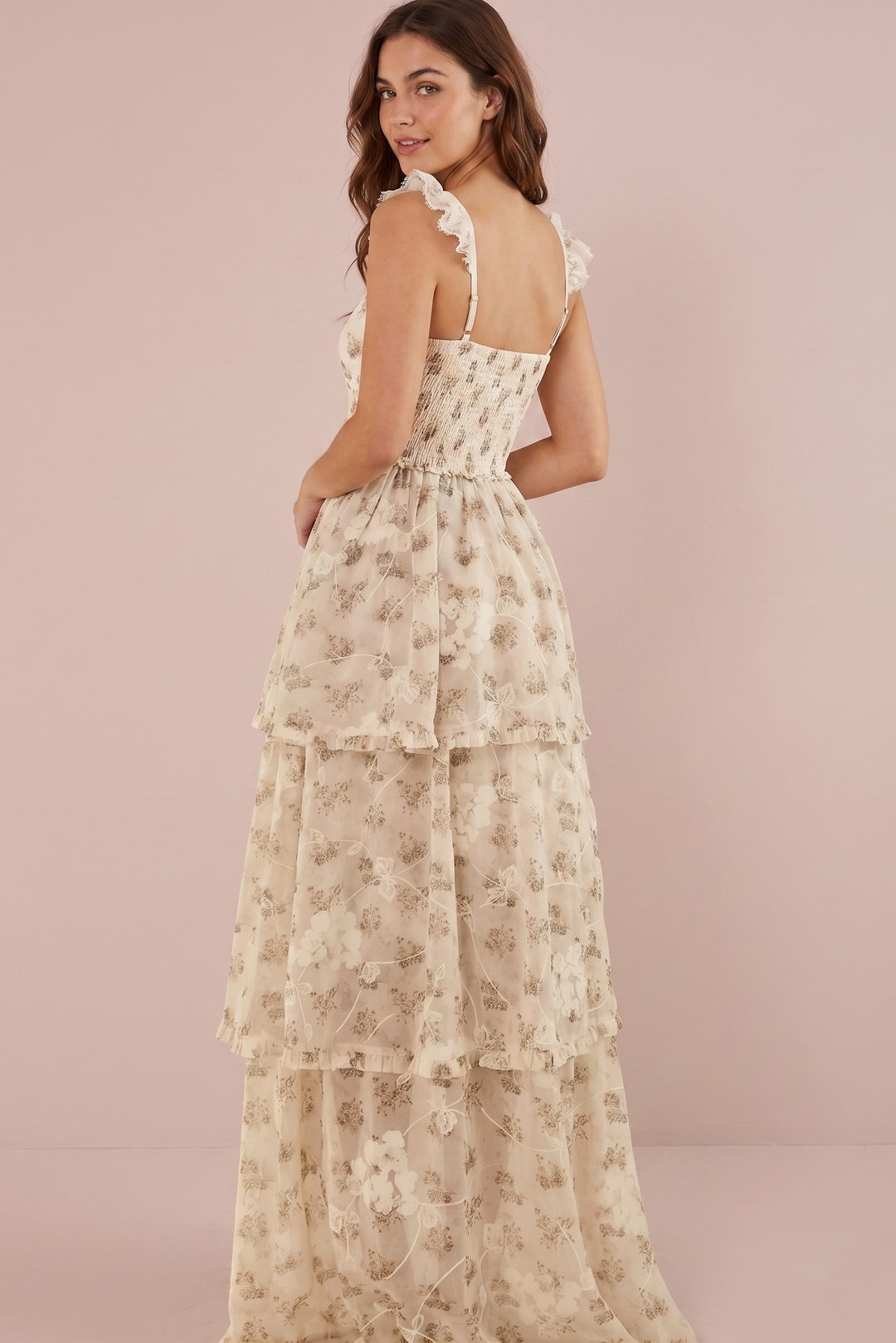 A woman modeling a cream-colored, tiered maxi dress with a delicate floral pattern and ruffled straps, photographed against a solid pale pink studio backdrop.