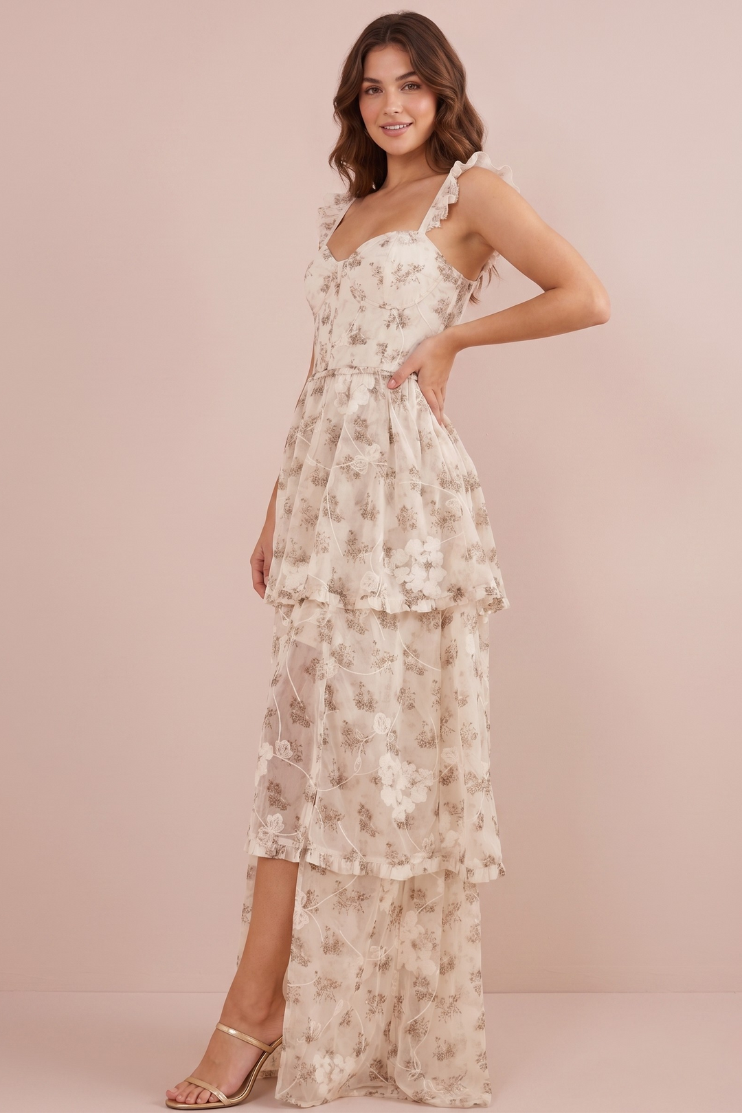 A woman models a long cream-colored tiered maxi dress with a delicate brown floral pattern, ruffled shoulder straps, and a high-low hemline, set against a solid pale pink studio backdrop.