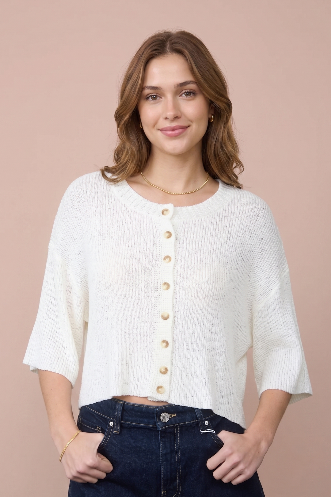 A woman wearing a cream-colored, short-sleeved button-up knit top and dark blue denim shorts, standing against a solid dusty pink background.
