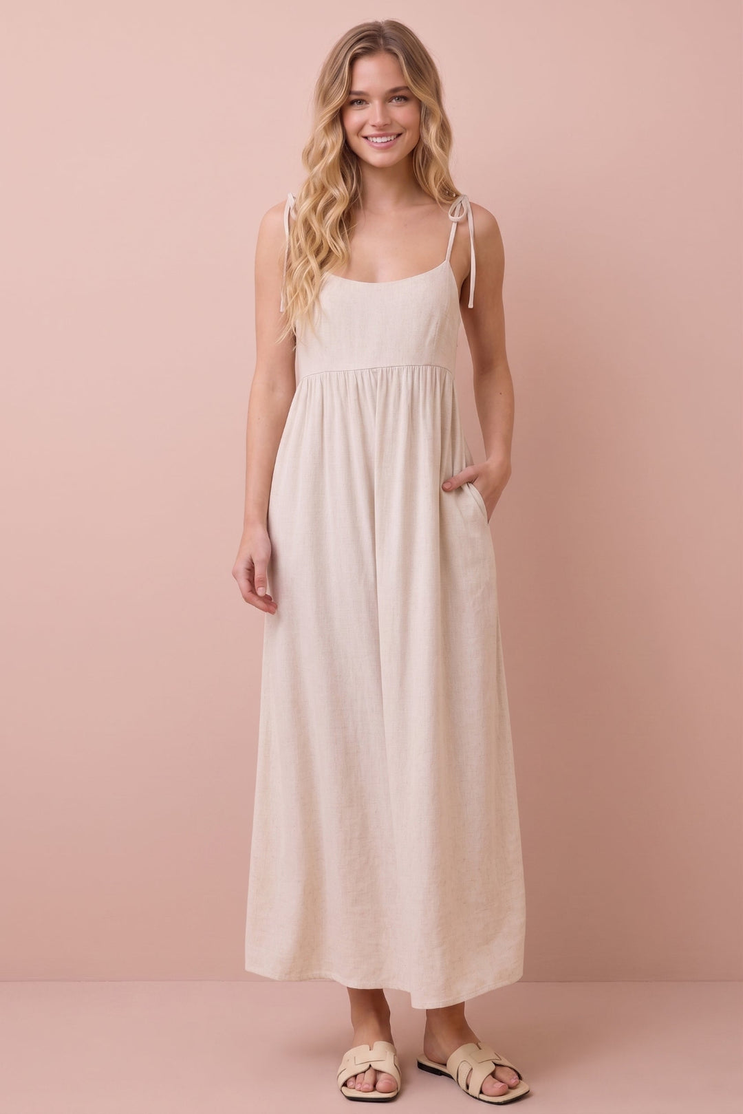 A woman modeling a light beige linen maxi dress with thin shoulder tie straps and side pockets, standing against a solid dusty pink background.