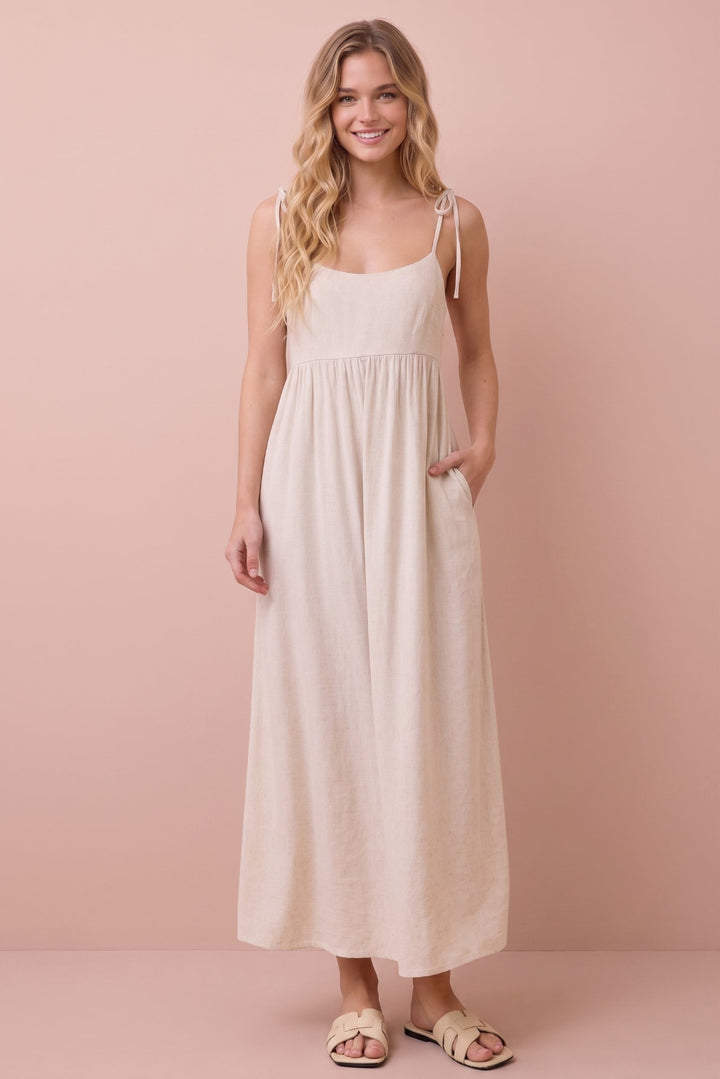 A woman modeling a light beige linen maxi dress with thin shoulder tie straps and side pockets, standing against a solid dusty pink background.