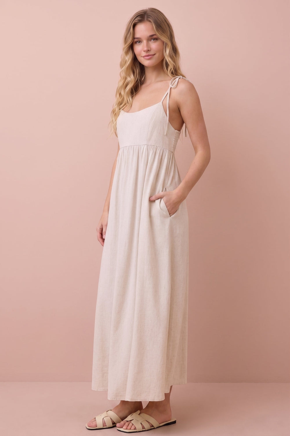 A woman modeling a light beige linen maxi dress with thin shoulder straps, side pockets, and a gathered waist, set against a solid dusty pink studio background.